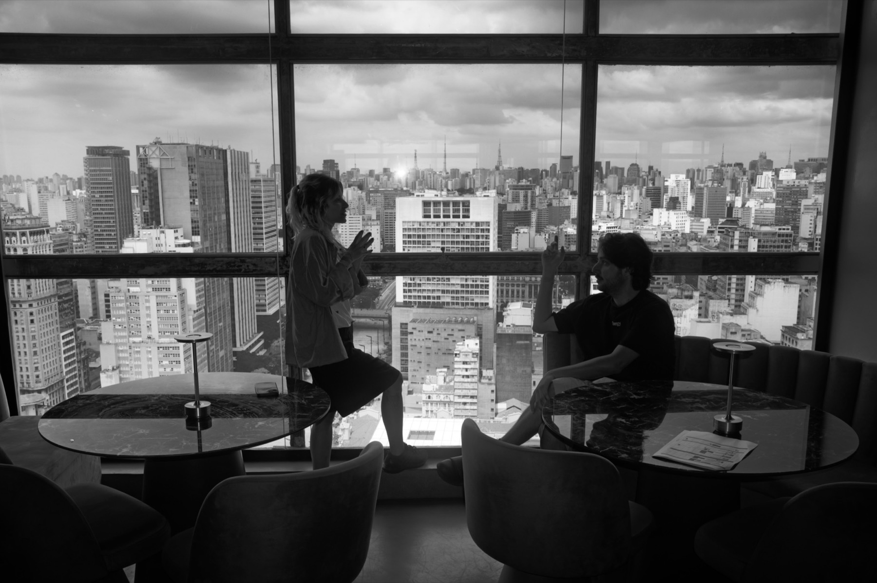 Two people in conversation by a full-height window over the São Paulo skyline